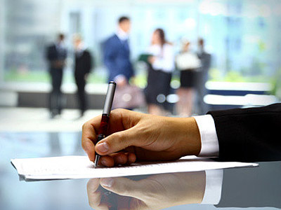 Eine Hand mit einem Stift, die auf einem Dokument schreibt, im Hintergrund sind Menschen in formeller Kleidung zu sehen.